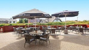 Un espace extérieur aménagé en terrasse, avec des tables rondes et des chaises en osier, ombragé par des parasols portant l’inscription « DE JYSKE BRYGHUSE ».