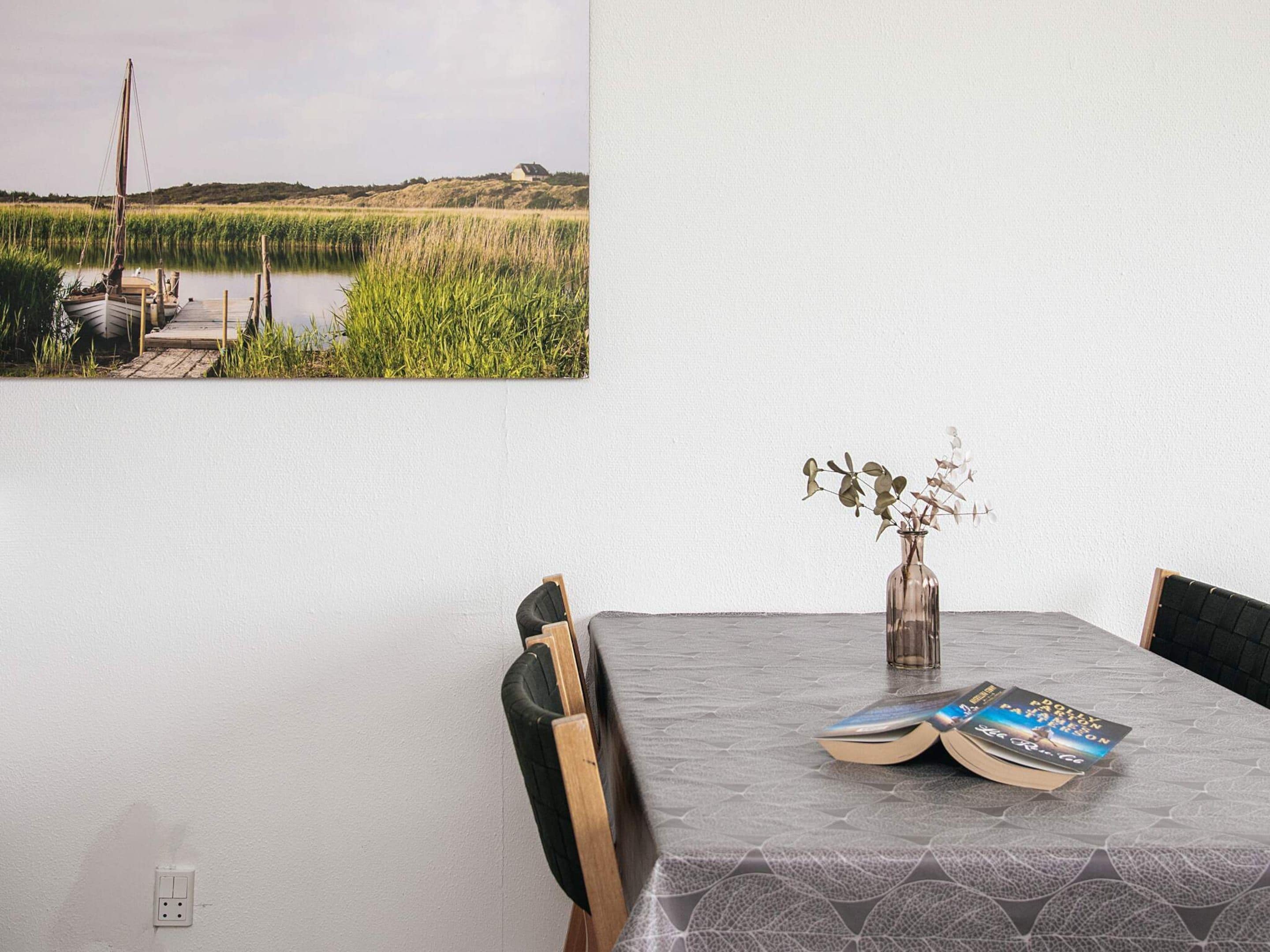 A dining table with a vase of dried flowers and books, a chair, and a wall-mounted painting of a landscape.