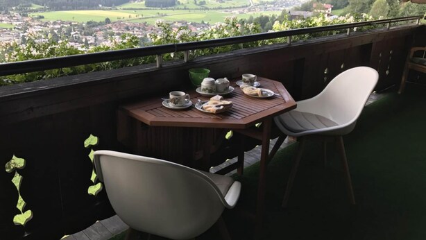 Un balcon avec une table préparée pour le thé, donnant sur une vallée parsemée de maisons et bordée de montagnes.