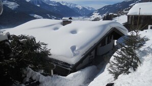 Un chalet de montagne enneigé, avec un toit en pente, entouré d’arbres couverts de neige et avec des montagnes enneigées en arrière-plan.