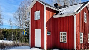 Une maison en bois rouge, avec une porte et des fenêtres blanches, entourée de neige et d’arbres.