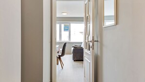 Un couloir mène à un espace de vie comprenant un canapé et des fauteuils, une fenêtre offrant une vue agréable et une porte dotée d’un miroir.