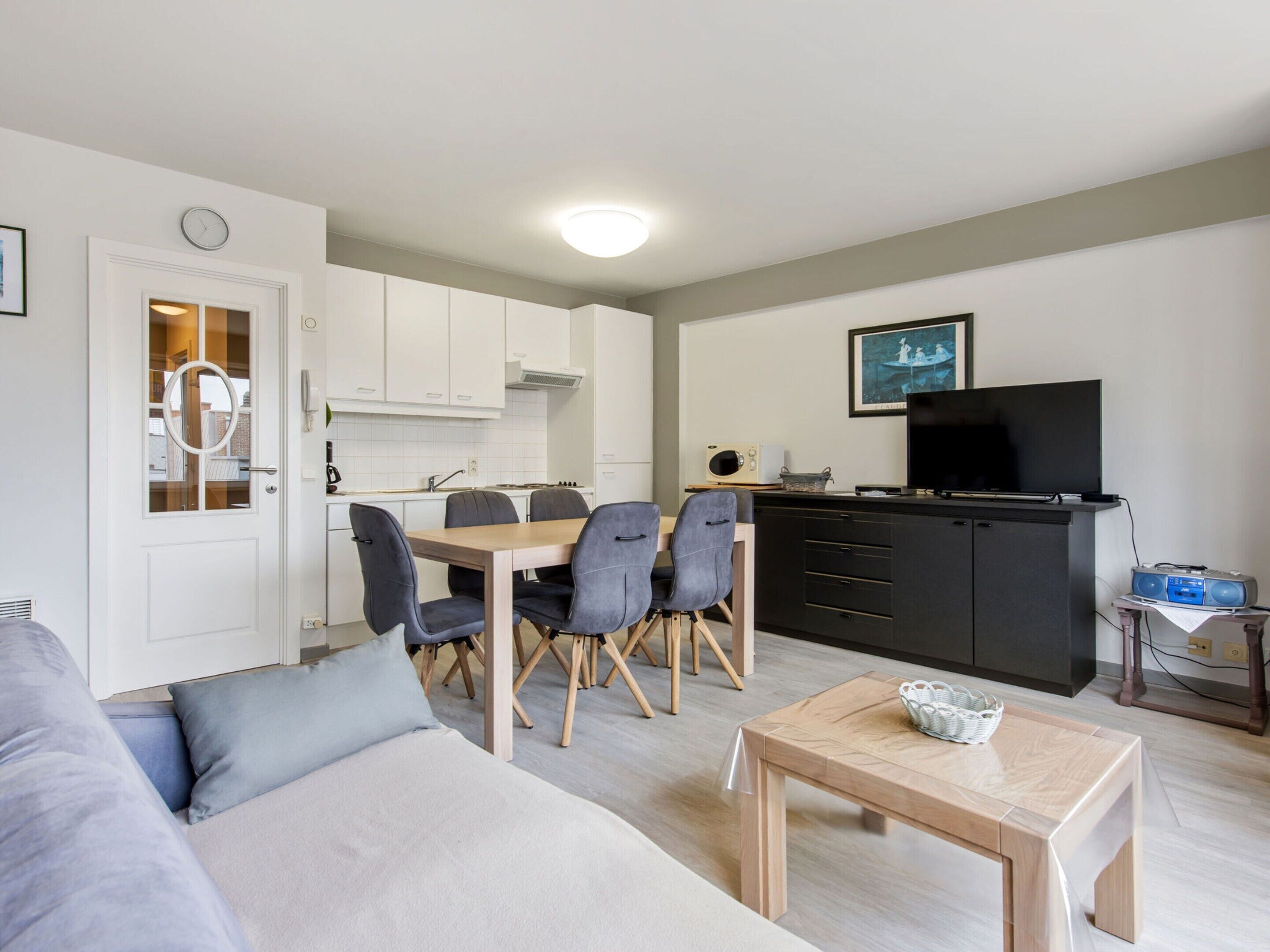 Una sala moderna con un sofá, una mesa de comedor con sillas, una zona de cocina con armarios, un televisor y una mesita con una cesta.