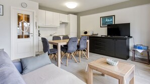 Un salon moderne comprenant un canapé, une table à manger avec des chaises, un coin cuisine avec des placards, un téléviseur et une petite table avec un panier.