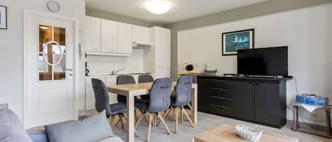 Una sala moderna con un sofá, una mesa de comedor con sillas, una zona de cocina con armarios, un televisor y una mesita con una cesta.