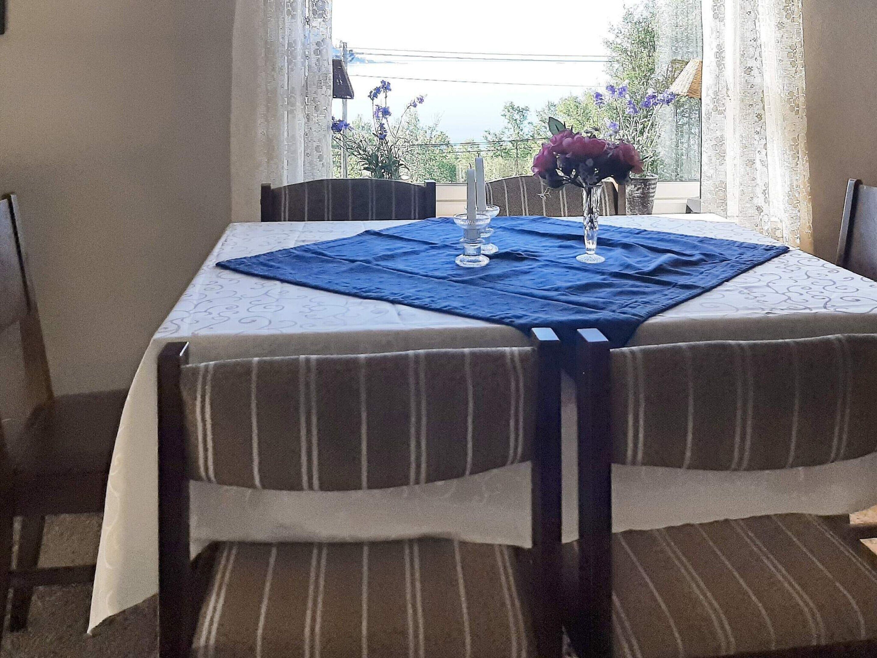 Une table à manger dressée avec un chemin de table bleu, une nappe blanche et deux vases en verre transparent contenant des fleurs violettes.