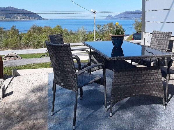 An outdoor dining area with wicker chairs and a table, overlooking a body of water and hills.