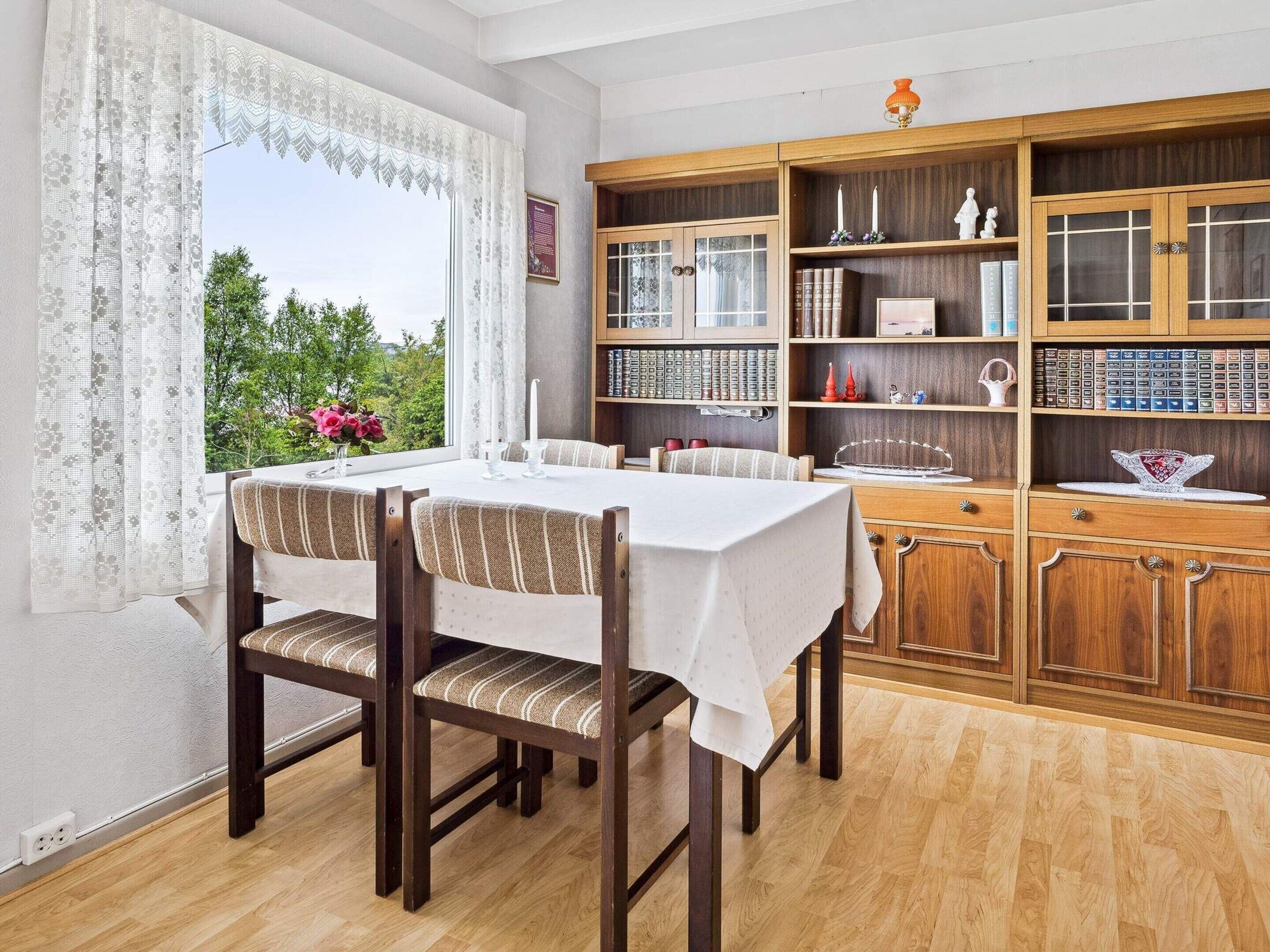A dining area with a table set for four, wooden chairs, a bookshelf with various items, and a window with curtains.
