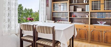 A dining area with a table set for four, wooden chairs, a bookshelf with various items, and a window with curtains.