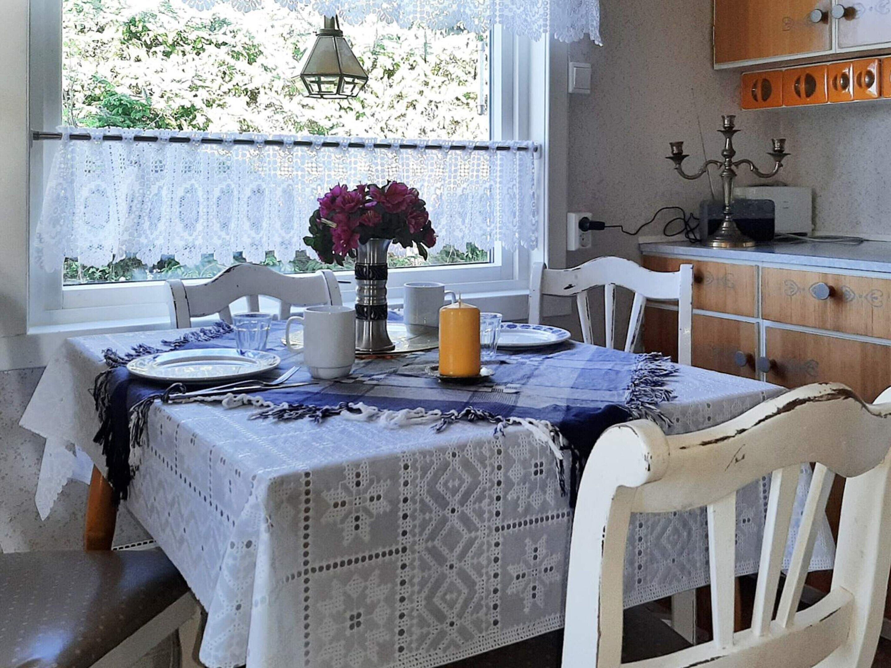 A dining area with a table set for two, a vase with flowers, and a view of greenery outside.