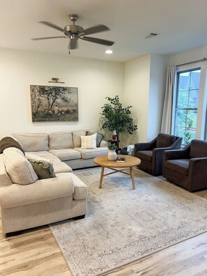 A living room with a sofa, armchairs, a coffee table, a ceiling fan, a painting, and a window.