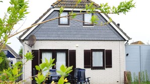 A two-story house with a grey tiled roof, white brick walls, and dark shutters. There is a satellite dish on the roof, and a blue chair is visible in the front yard. - Huijsmans Verhuur Type B de Luxe Zuiderdiep 8 (Ouddorp)