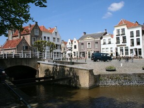 Uma praça histórica da cidade com uma ponte sobre um canal, rodeada por edifícios europeus tradicionais.