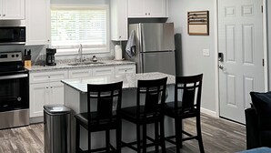 A modern kitchen with white cabinets, a stainless steel refrigerator, a coffee maker, and a trash can. There are three black chairs around a small island. - Cozy Cabin at Villager Resort on Lake Cumberland KY (Somerset)