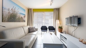 A modern living room with a sofa, two chairs, a dining table, a TV, and a painting of a beach scene. - Cosy Apartment With a Terrace (Middelkerke)