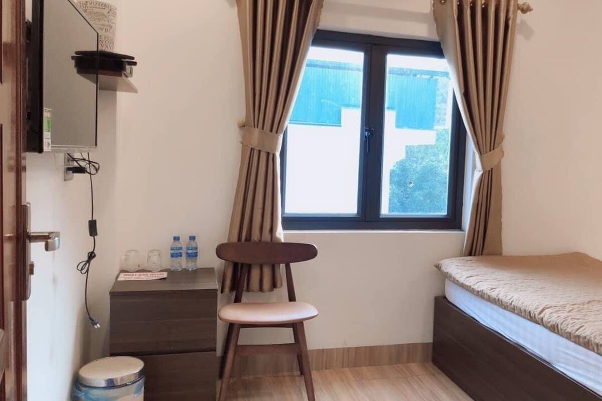 A small, tidy room with a single bed, a chair, a small table with a bottle, and a window with curtains.