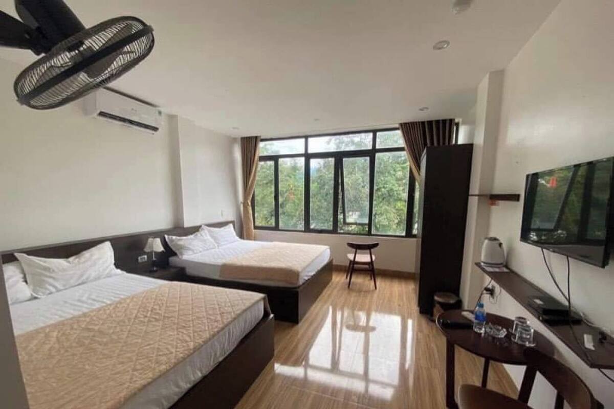 A hotel room with two beds, a desk with a chair, a TV, and a window with a view of trees.