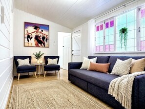 A living room with a dark blue sofa, two armchairs, a woven rug, a painting on the wall, and a window with a plant.