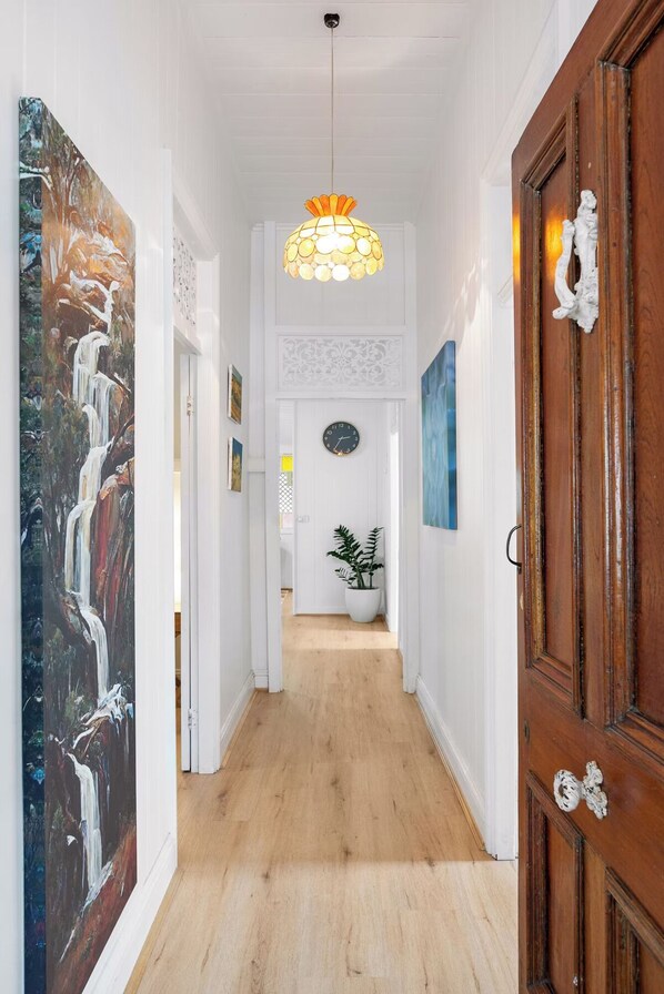 A hallway with a wooden floor, white walls, a large painting on the left, a potted plant in the center, and a wooden door on the right.