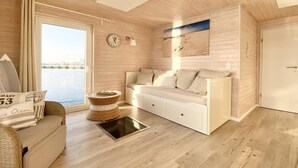 A room with a wooden floor, a white sofa, a round wicker side table, a clock, and a view of the sea through a large window. - Houseboat South Beach (Fehmarn)