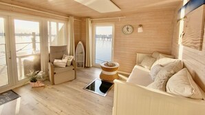 A cozy cabin with a wooden interior, a sofa, a chair, a surfboard, and a clock on the wall. - Houseboat South Beach (Fehmarn)