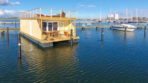 A floating structure with a covered deck and a small enclosed area, surrounded by calm water and anchored by poles. - Houseboat South Beach (Fehmarn)