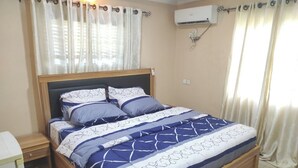 A bedroom with a wooden bed, a bedside table, a nightstand, a window with sheer curtains, and an air conditioning unit on the wall. - Abeokuta 2-Bed House, Sleeps 4 with Free Parking (Abeokuta)