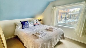 Cama bem arrumada com colcha branca e cinza, duas almofadas com motivos náuticos e janela com vista para área externa com mesas e cadeiras.