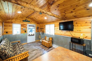 A cabin interior with a wooden ceiling, a television mounted on the wall, a sofa, a small table, and a microwave. - Tiny cabin in towering pines. Pets accepted! (Nevis)