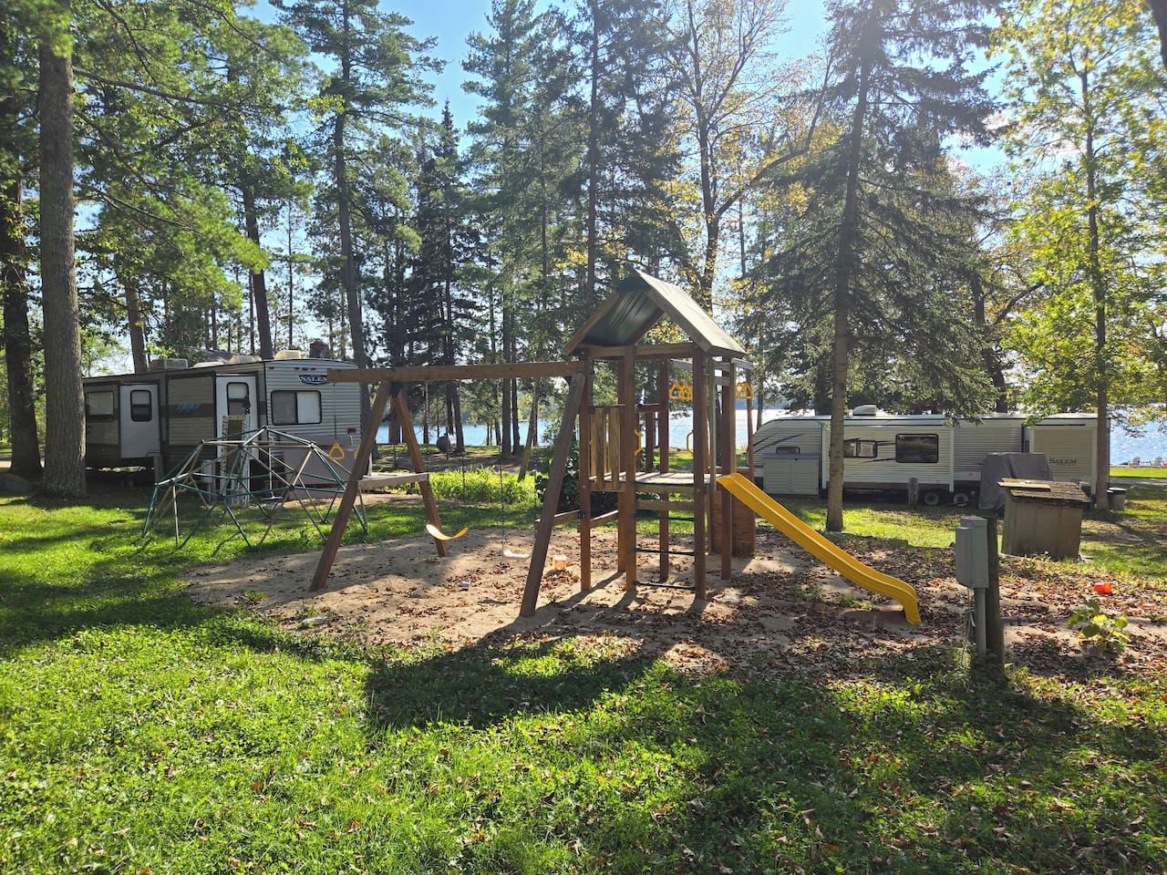 A playground with swings and a slide, surrounded by trees and RVs.