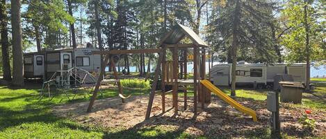 A playground with swings and a slide, surrounded by trees and RVs.