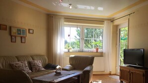 A living room with a sofa, a coffee table, a television on a wooden cabinet, and a window with curtains. - Holiday Apartment Haus Kirsch (Hilders)