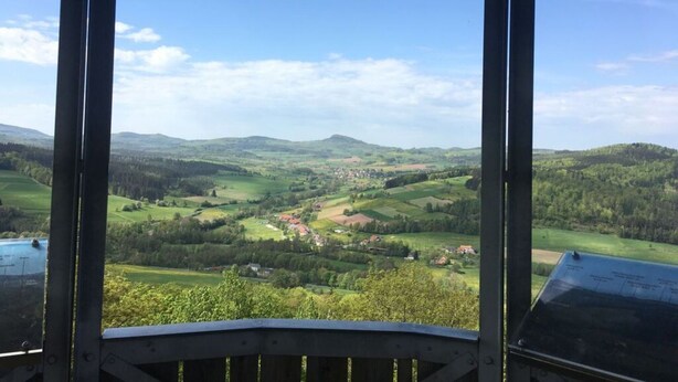 A view from a covered area with a wooden railing, overlooking a valley with scattered houses and green fields under a clear sky. - Holiday Apartment Haus Kirsch (Hilders)