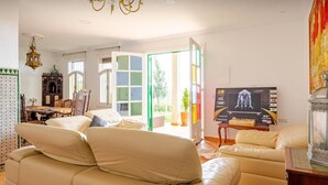 Een ruime woonkamer met een grote beige bank, een televisie waarop een filmaffiche staat en een kroonluchter.