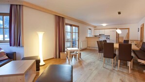 A modern living room with a sofa, a dining table with chairs, a large window with curtains, and a kitchen area in the background. - Apartment in Wald im Pinzgau Near the ski Lifts (Wald im Pinzgau)
