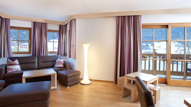 A modern living room with a sofa, ottoman, and coffee table, large windows with curtains, and a view of a snowy landscape. - Apartment in Wald im Pinzgau Near the ski Lifts (Wald im Pinzgau)