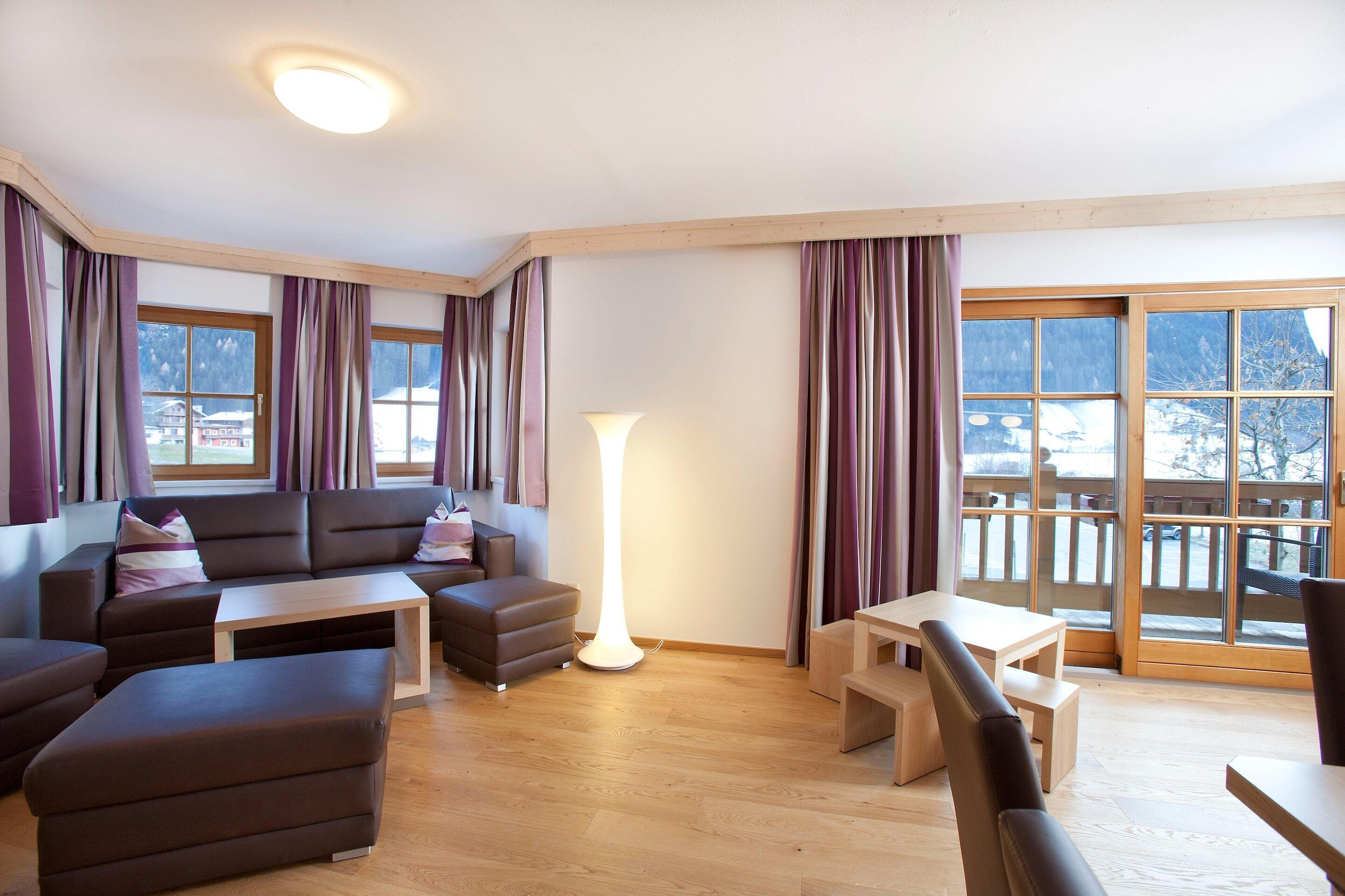 Ein modernes Wohnzimmer mit einer Couch, Hocker und einem Tisch, großen Fenstern mit Vorhängen und Blick auf verschneite Berge.