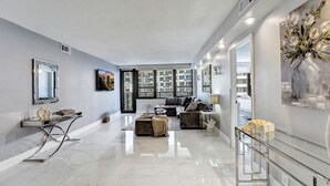 A modern living room with a sofa, ottoman, and a glass coffee table. There is a painting on the wall and a mirror above the coffee table. - Amazing Ocean Front Condo / 2BD beach Resort / 1515 (Miami Beach)