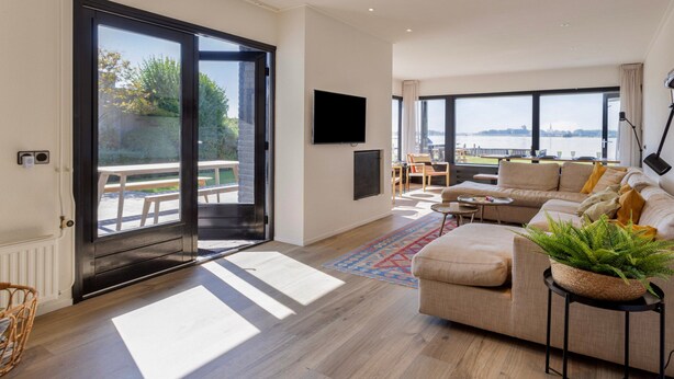 A modern living room with a large sliding glass door leading to an outdoor area, a sofa, a coffee table, and a television mounted on the wall. - Lakeside Stay in Kamperland (Kamperland)