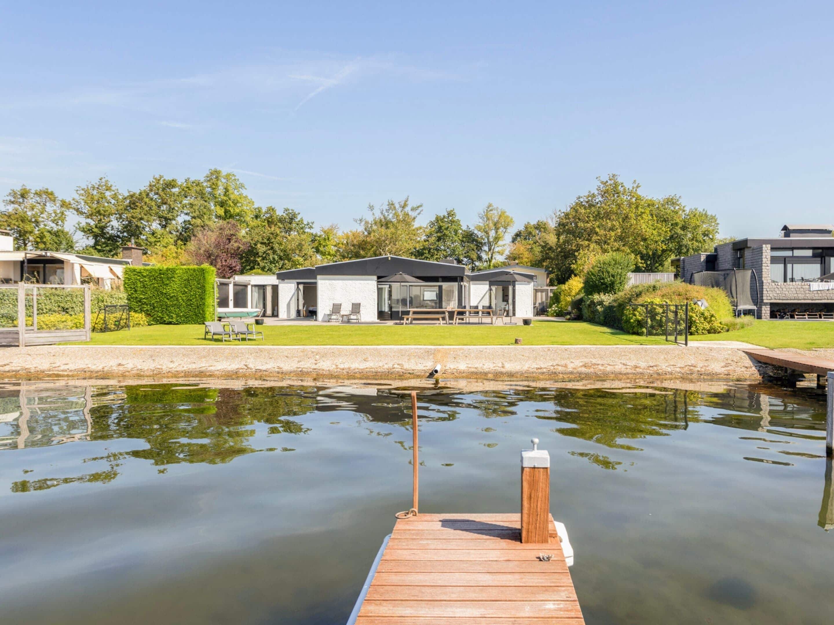 Una casa moderna con un muelle que se adentra en un lago tranquilo, rodeada de abundante vegetación y otras viviendas.
