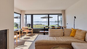 A modern living room with a large window overlooking a body of water, a sofa, a coffee table, and a dining area with a table and chairs. - Lakeside Stay in Kamperland (Kamperland)