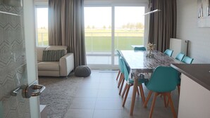 A modern living room with a sofa, a dining table with chairs, and a view of a green field through large windows. - Apartment in Nieuwpoort Near the Marina (Nieuwpoort)