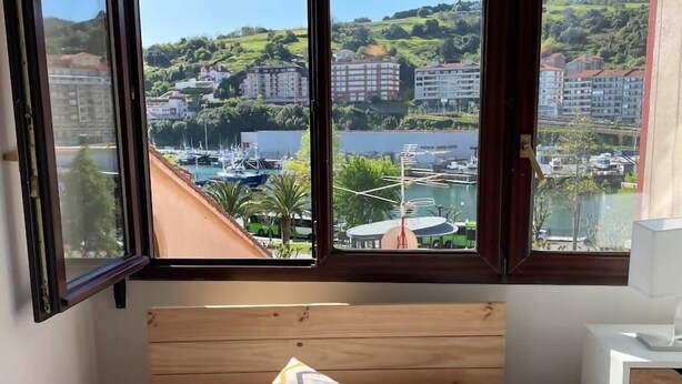 A bedroom with a wooden headboard, a bed with white bedding, and a view of a marina and buildings through large windows. - Bermeo Park - Centrica Casa Independiente (Bermeo)