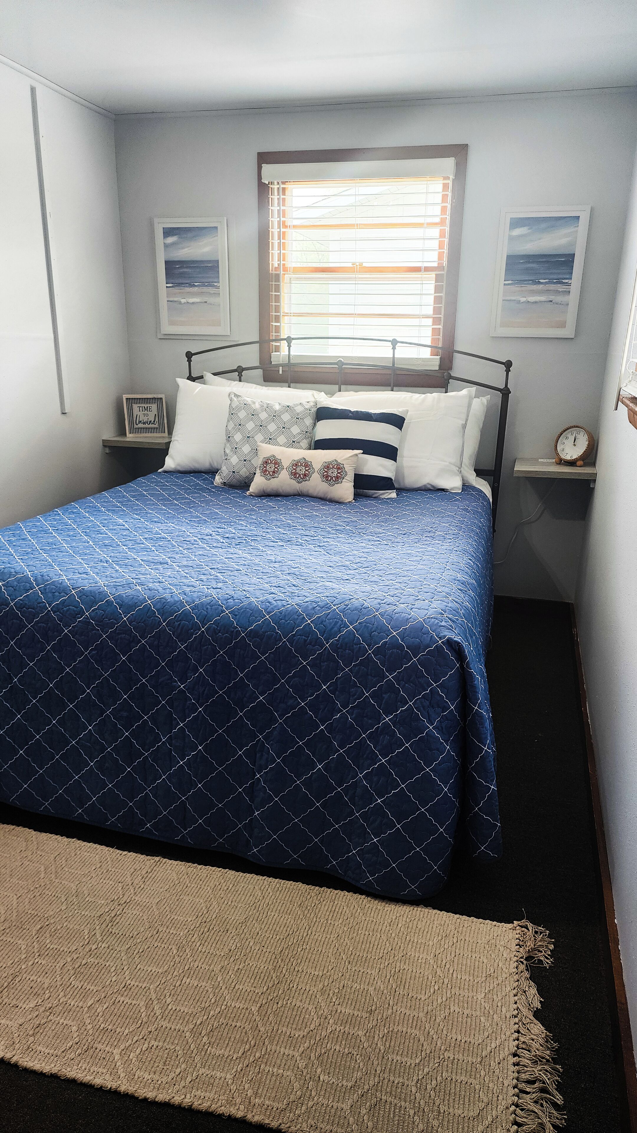 A bedroom with a bed, a window with blinds, a clock, and two framed pictures on the wall.