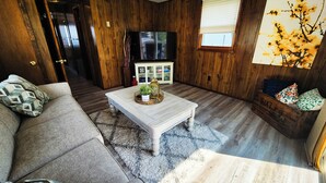 Una sala de estar acogedora con suelo de madera, una mesa de centro blanca con una planta, un sofá gris y un aparador de madera al fondo.