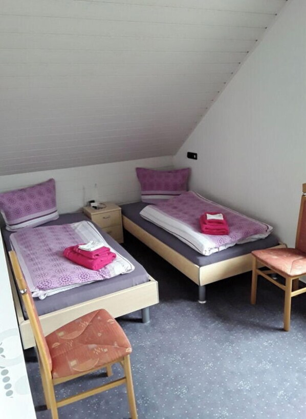 A small, single-room accommodation with two beds, a desk, and a chair.