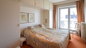 Een slaapkamer met een bed, een klein krukje, een radiator en een raam met gordijnen.