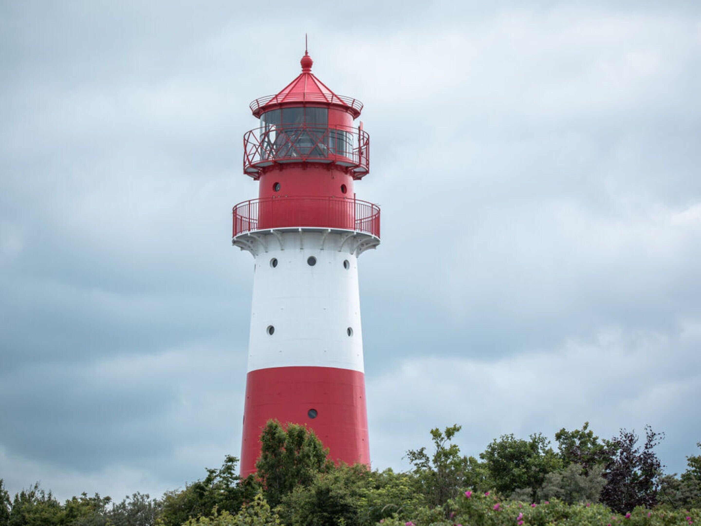 A tall lighthouse with a red top and white lower section, surrounded by greenery.