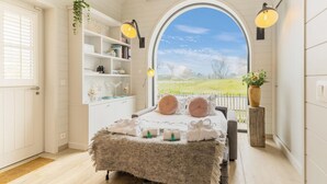 Une chambre dotée d’une grande fenêtre en arc de cercle, d’un lit recouvert d’une couverture moelleuse, de lampes de chevet et d’une étagère ornée d’objets décoratifs.