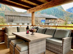Un espace extérieur aménagé, abrité et doté de mobilier en osier et d’une table, offrant une vue sur un paysage montagneux parsemé de bâtiments traditionnels.